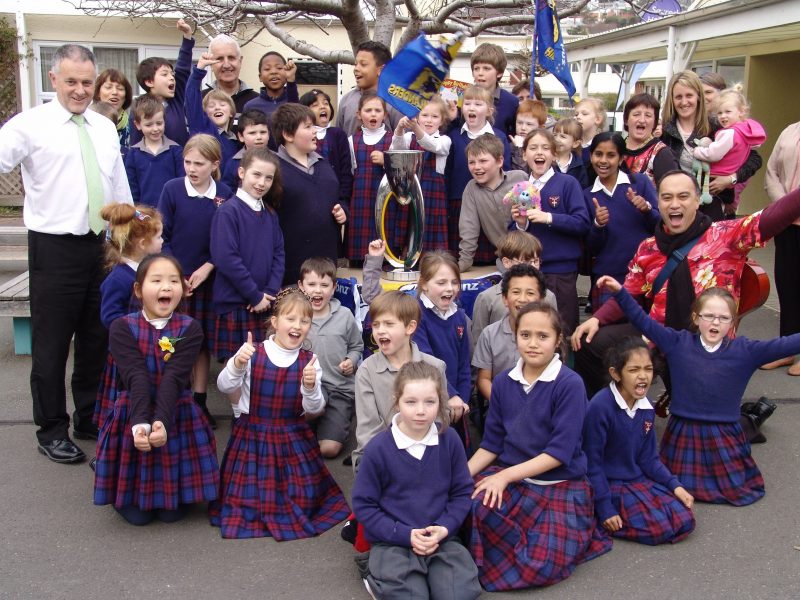 Sacred Heart pupils show their delight in learning about their rugby franchise’s trophy. Unobscured adults, from left to right, principal Paul Richardson, teacher Kath Rutherford, grandparent Michael Botting, teacher Bronwyn Bradshaw, parent Emmeline Kendrick and (kneeling) DRS Pesamino-  Solomona Tili.